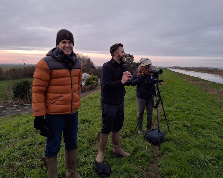 Three people standing on a grassy river bank, one with a telescope, counting swans in the early morning.